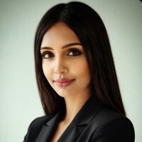 Woman with long straight dark hair, wearing a black blazer, poses against a plain light background, looking at the camera with a neutral expression.