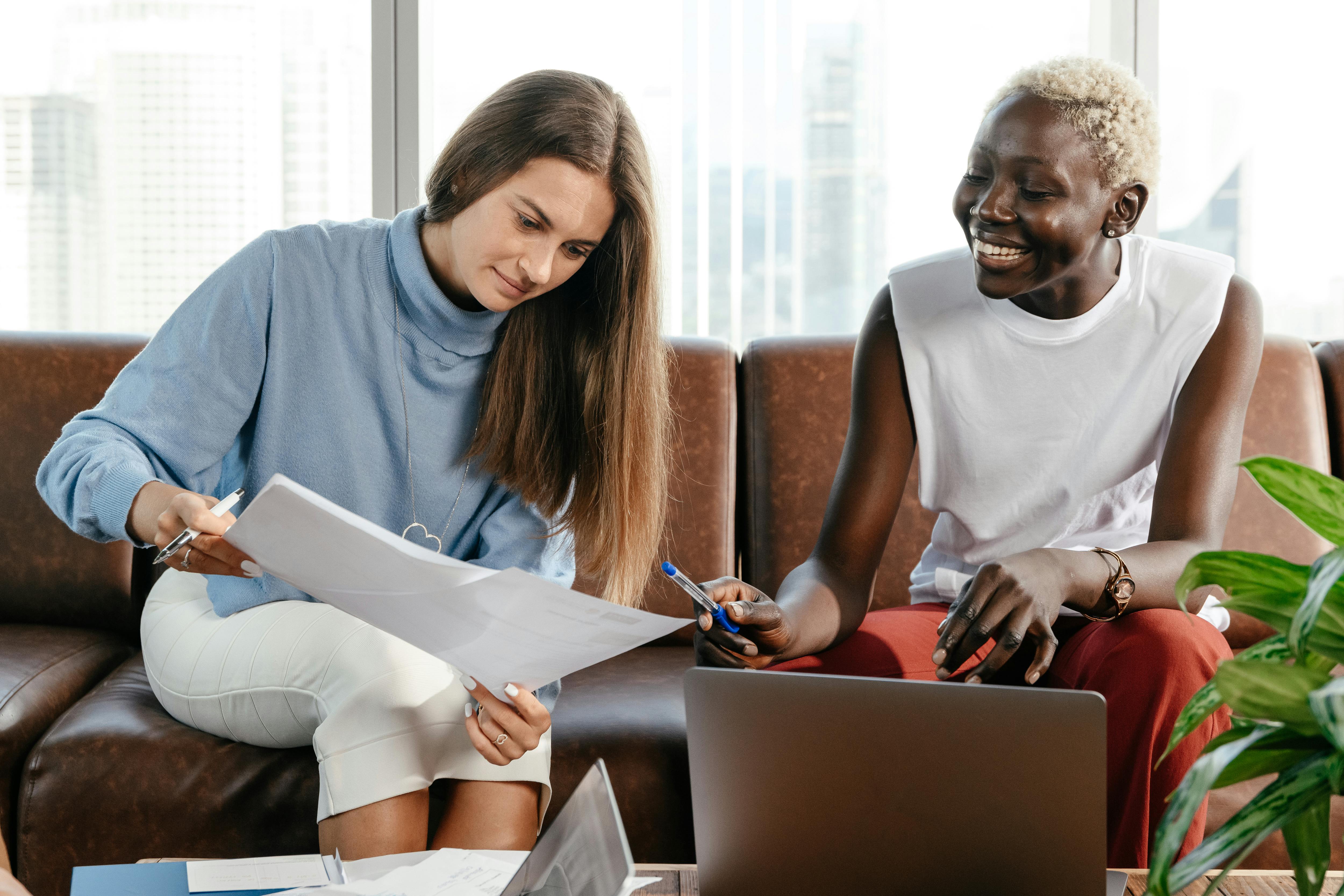 Two people sit on a couch reviewing documents together, one holding papers and a pen, the other smiling, with a laptop open in front of them.