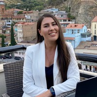 Woman with long brown hair wearing a white blazer sits at an outdoor table with a laptop, smiling, with a cityscape and hills in the background.
