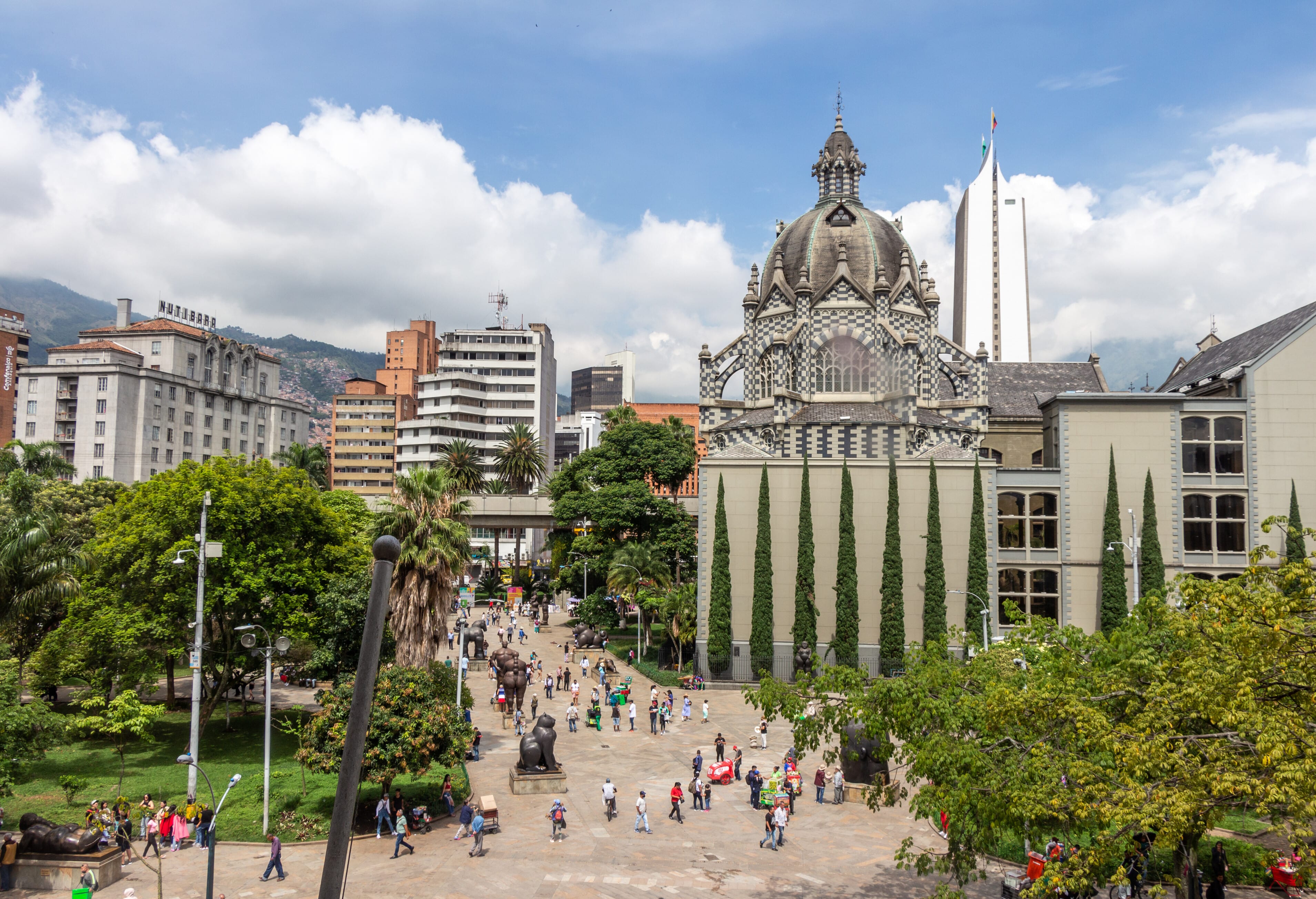 A busy public square with people walking, a patterned dome building, tall cypress trees, modern buildings, and mountains in the background under a partly cloudy sky.