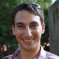 A man with short brown hair and sunglasses on his head smiles at the camera, wearing a plaid shirt, with greenery in the background.