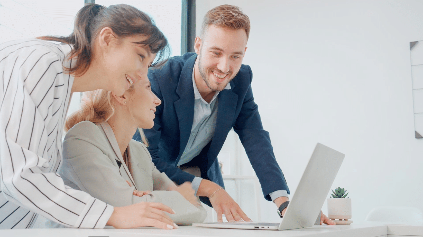 Three people smiling and looking at a laptop screen in an office setting, with a desk and a potted plant visible.