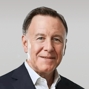A middle-aged man in a dark suit jacket and white collared shirt, smiling at the camera against a plain light grey background.