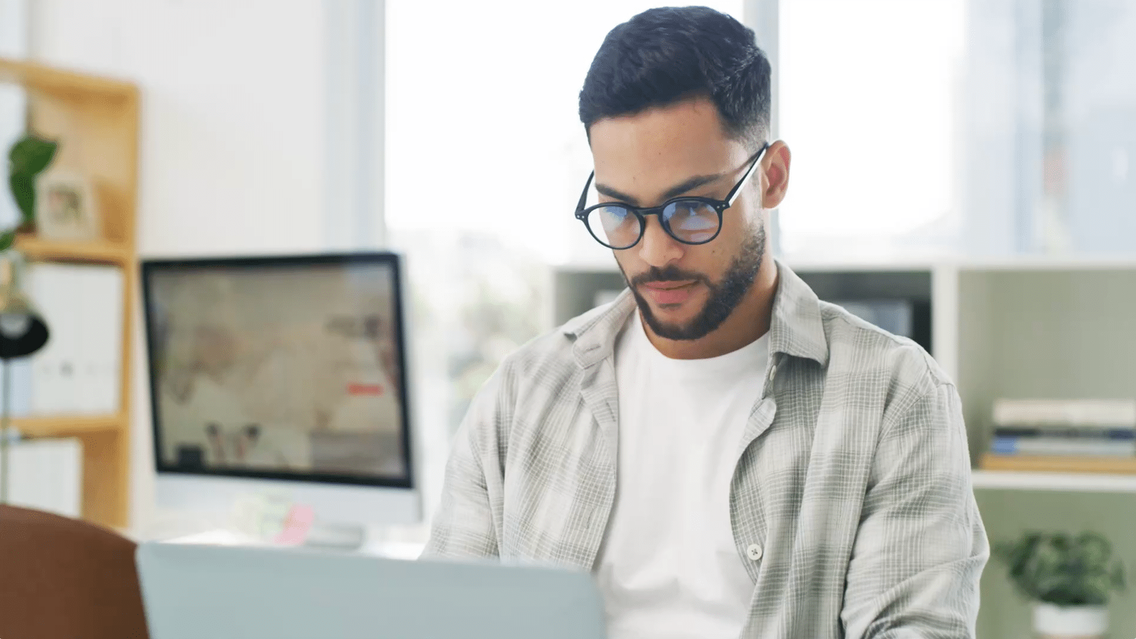 Man with glasses sitting and working on a laptop in a brightly lit room. A blurred background with a desk and shelves is visible.