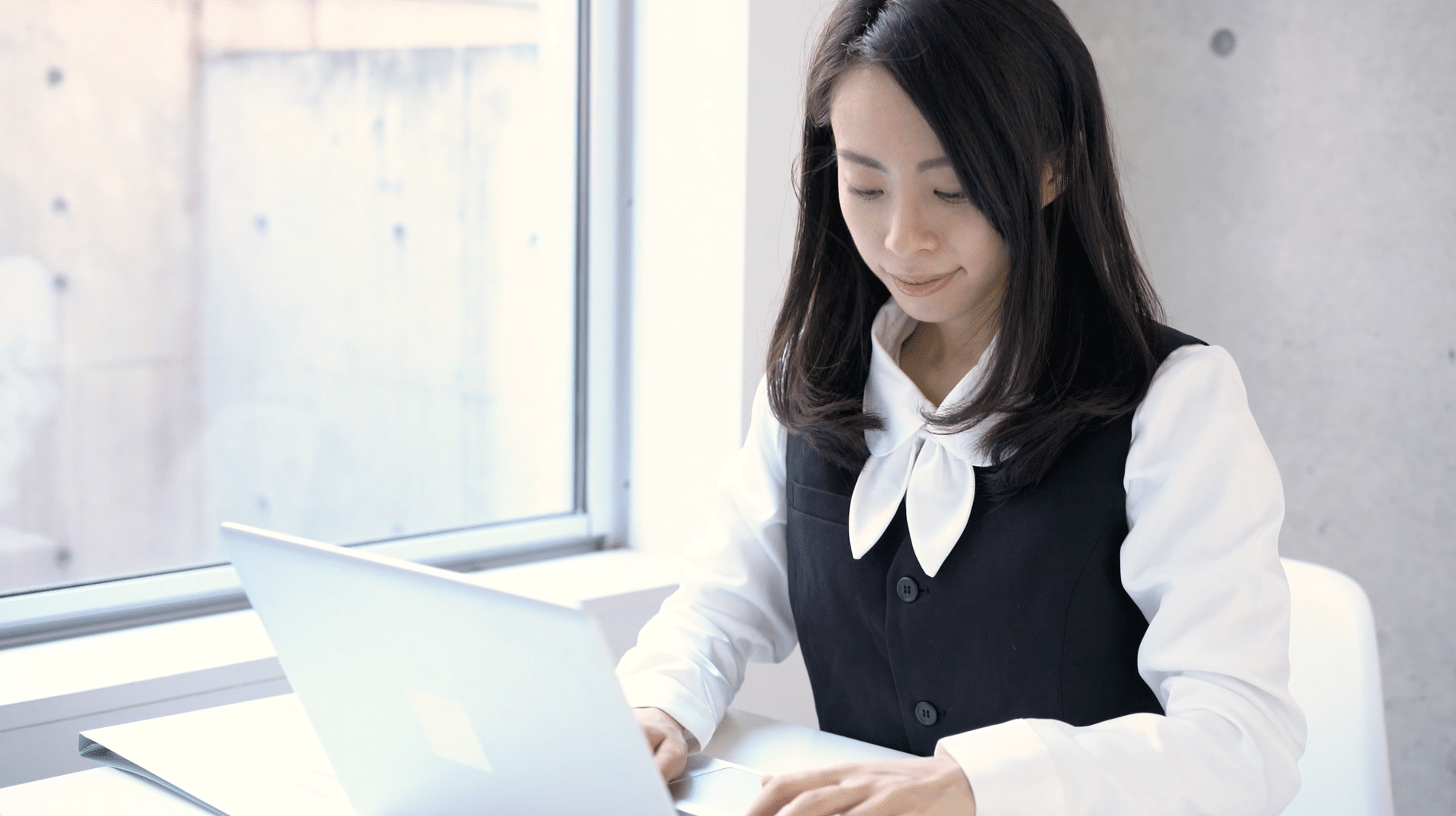 A person in a white blouse and black vest is working on a laptop by a window with soft natural light.