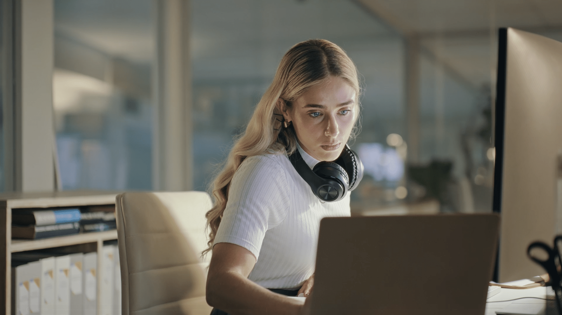 Woman with headphones around neck working on a laptop at a desk in an office.