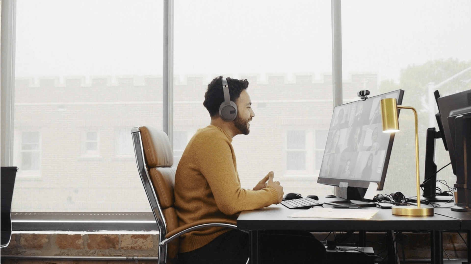 A man in a yellow sweater sits at a desk, wearing headphones, engaged in a video call on a computer in a bright office.