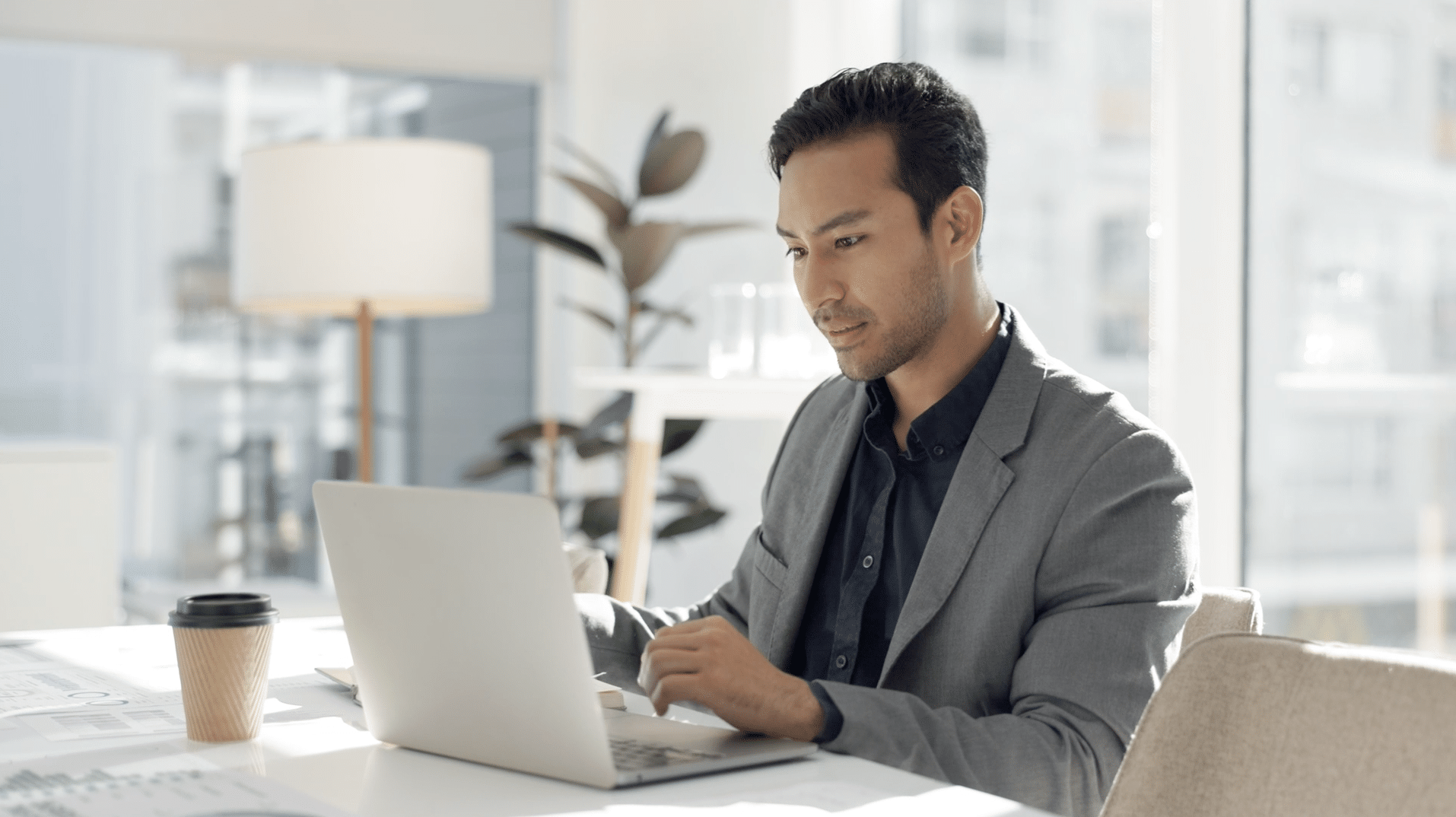 A man in a suit works on a laptop at a desk in a bright office. A plant, lamp, and coffee cup are in the background.