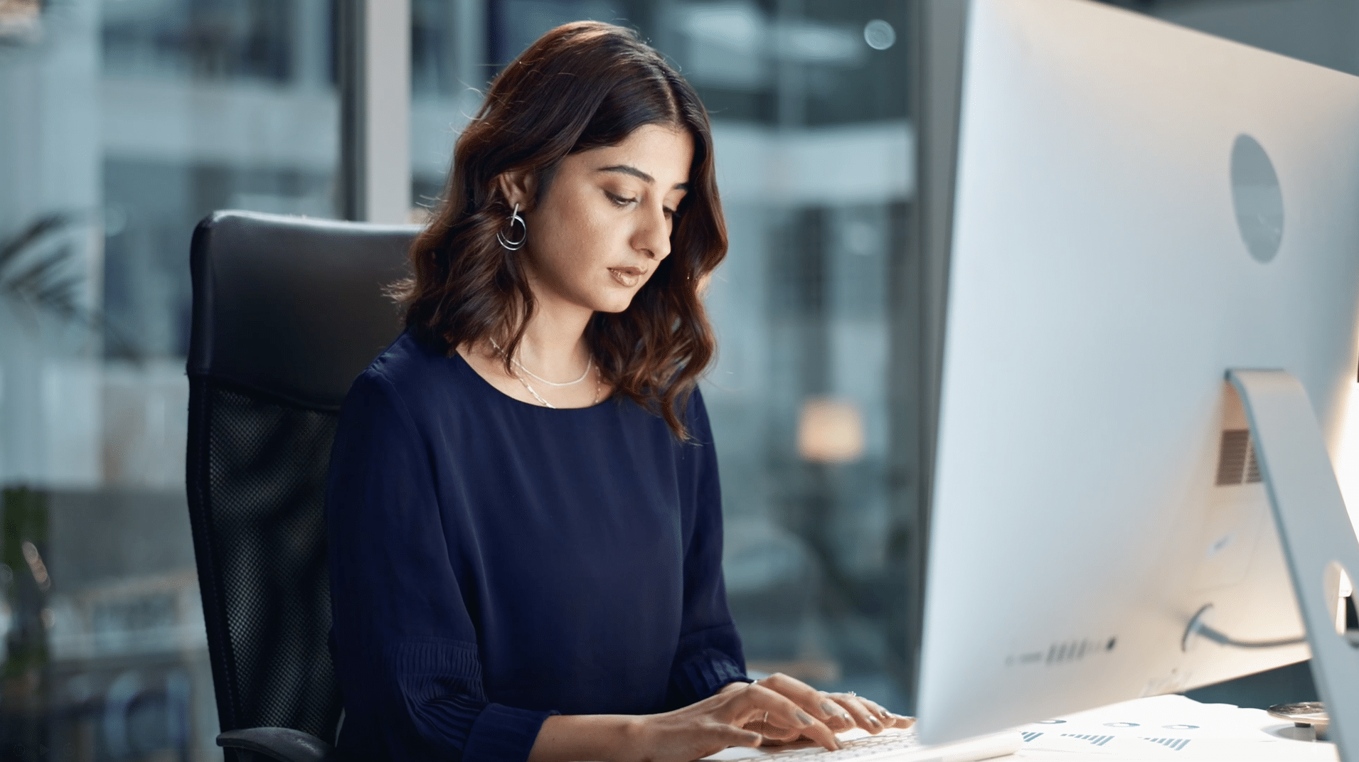 A woman in a navy blouse types on a keyboard while sitting at a desk with a computer monitor in a modern office setting.