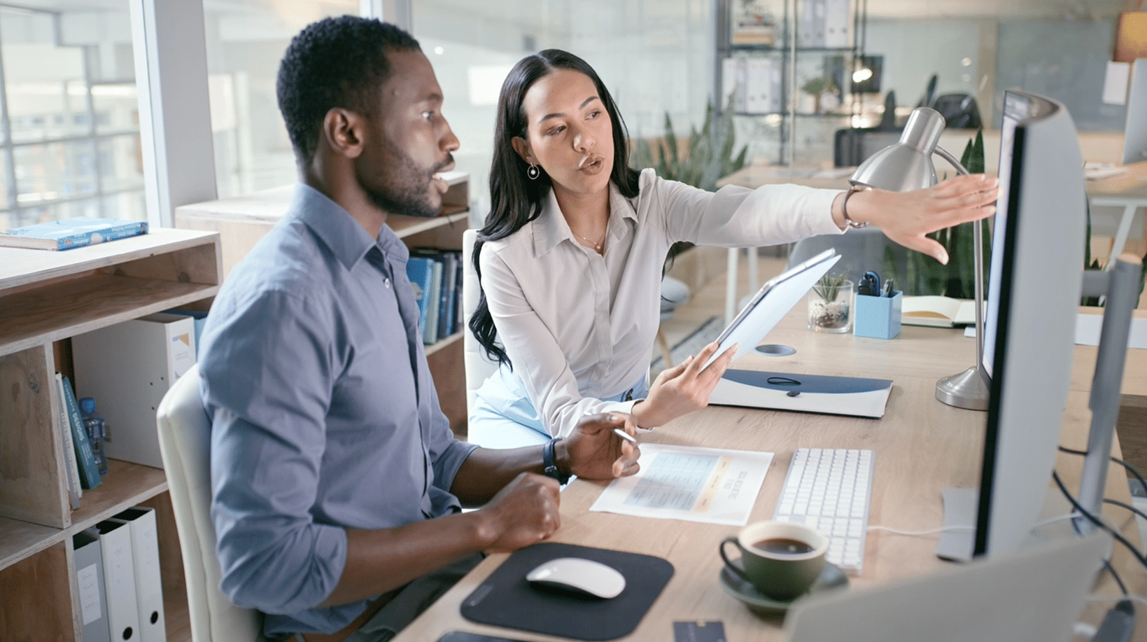 Two people at a desk, one holding papers and pointing at a computer screen, discussing work in an office setting.