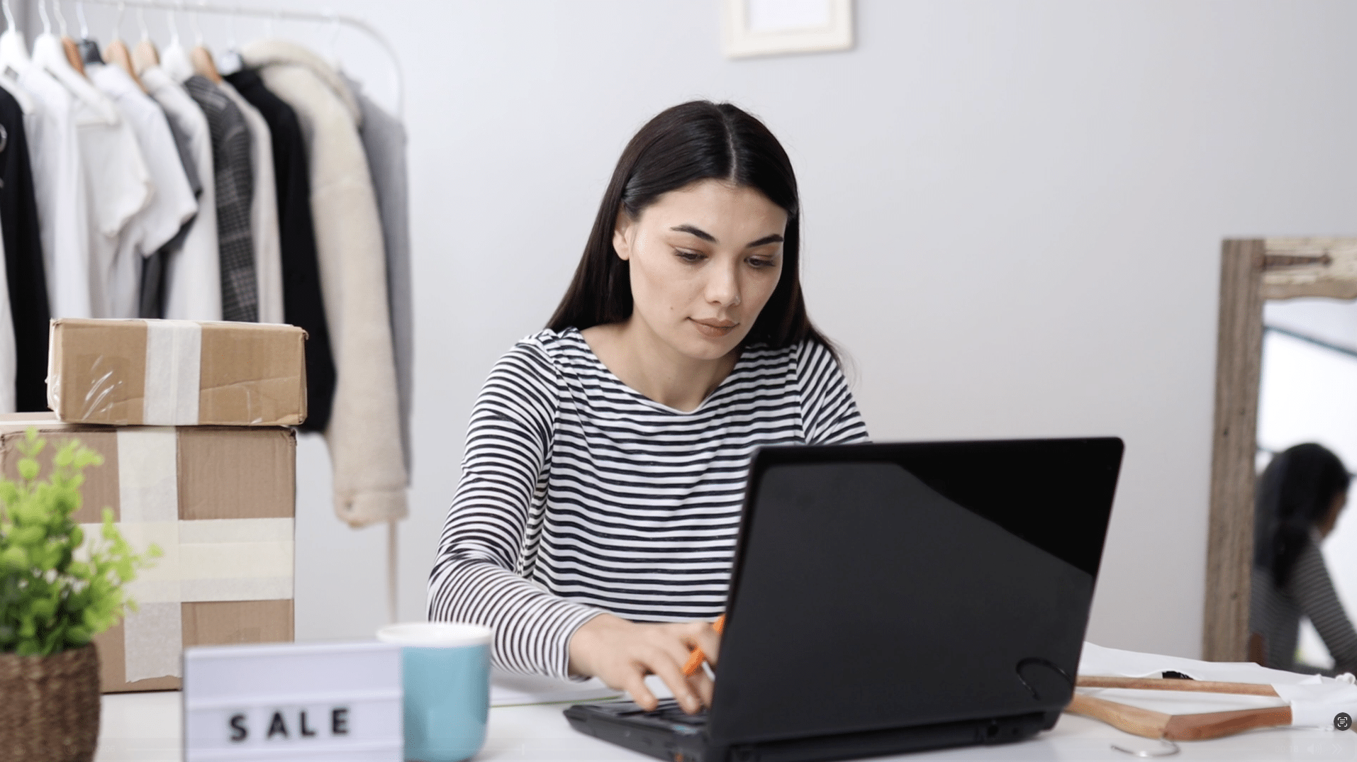 Woman in a striped shirt using a laptop at a desk with a plant, mug, and sale sign, with clothes and boxes in the background.