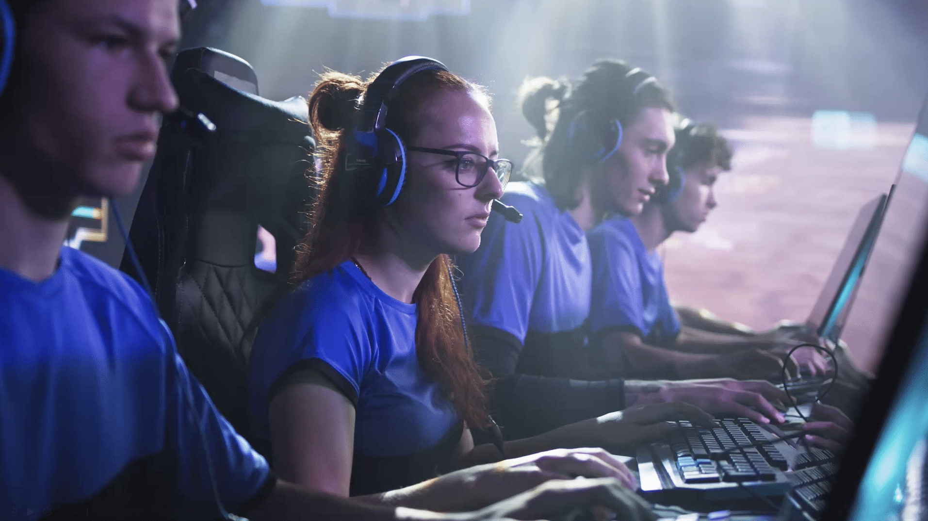 Three people wearing headsets focus on gaming screens, illuminated by blue light. They appear to be part of a team, dressed in matching blue and black shirts.