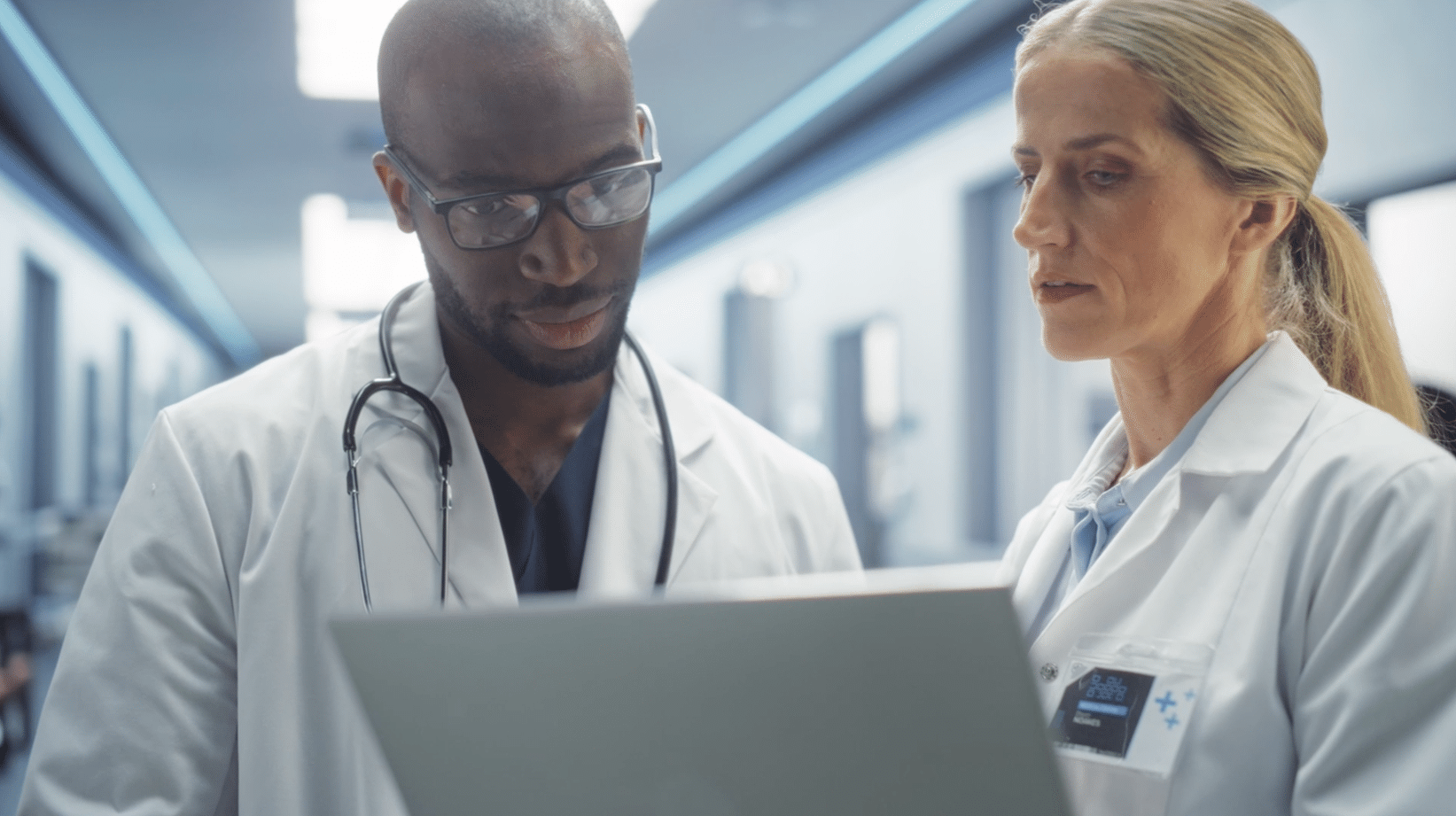 Two medical professionals in lab coats review information on a laptop in a hospital corridor.