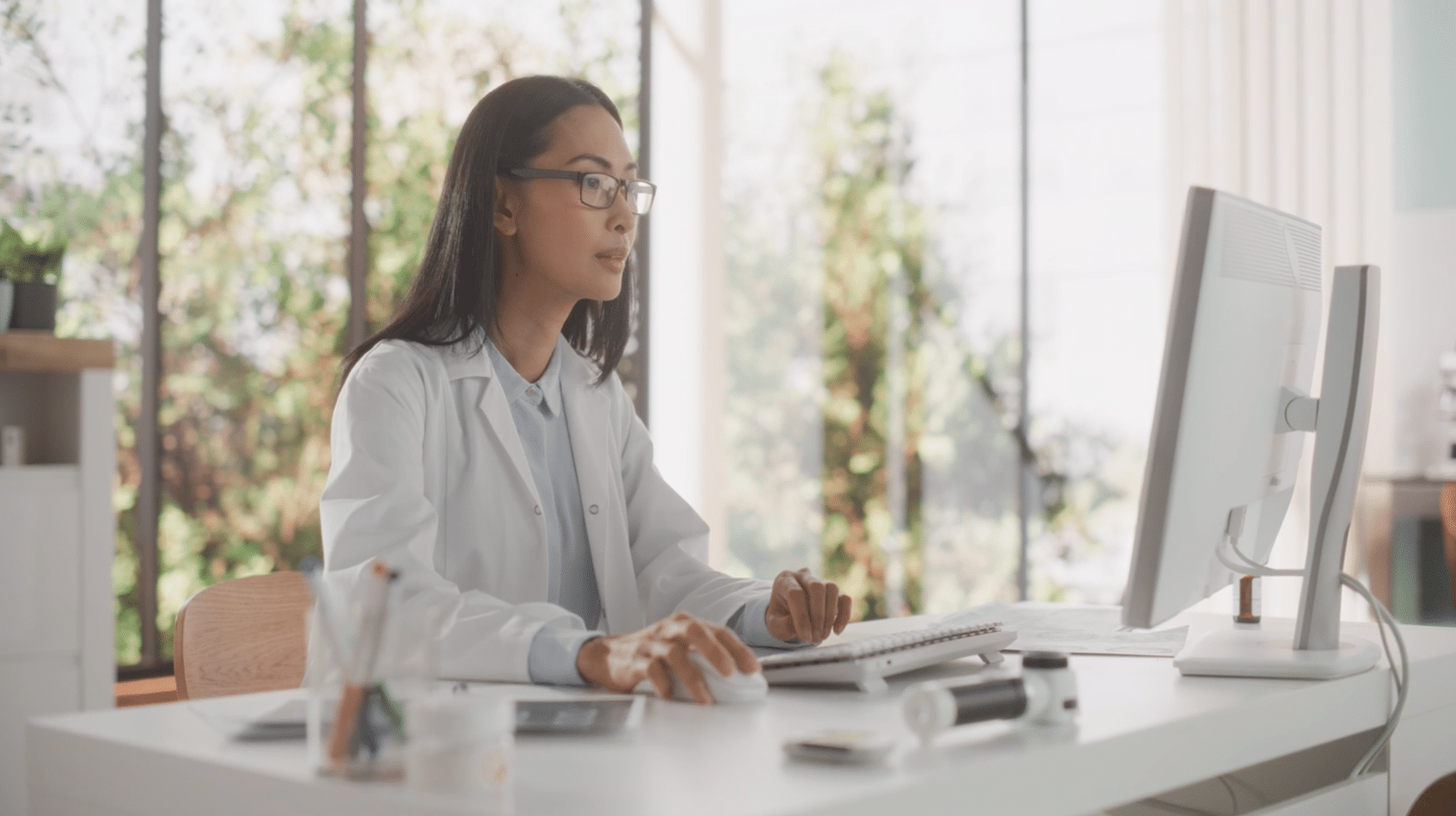 A person wearing glasses and a lab coat sits at a desk, working on a computer in a bright room with large windows.