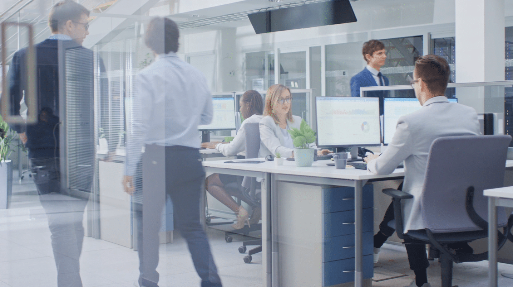 People working in a modern office with computers on desks, one person walking past. Bright, open space with glass partitions and visible office equipment.