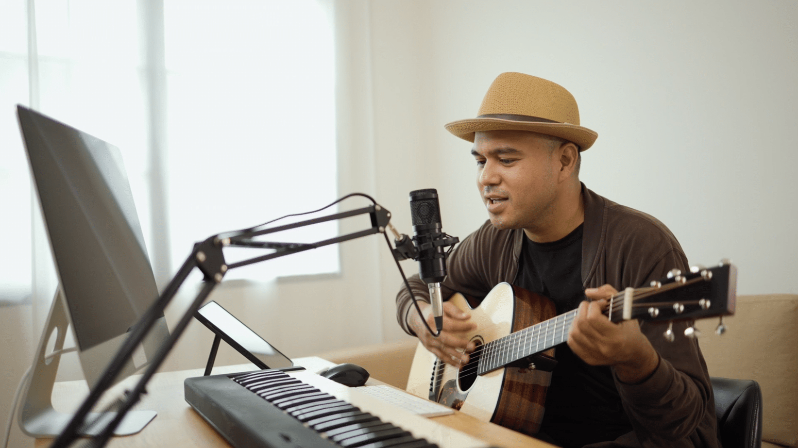 Person wearing a hat plays an acoustic guitar and sings into a microphone. There is a keyboard on the desk and a computer screen in front of them.