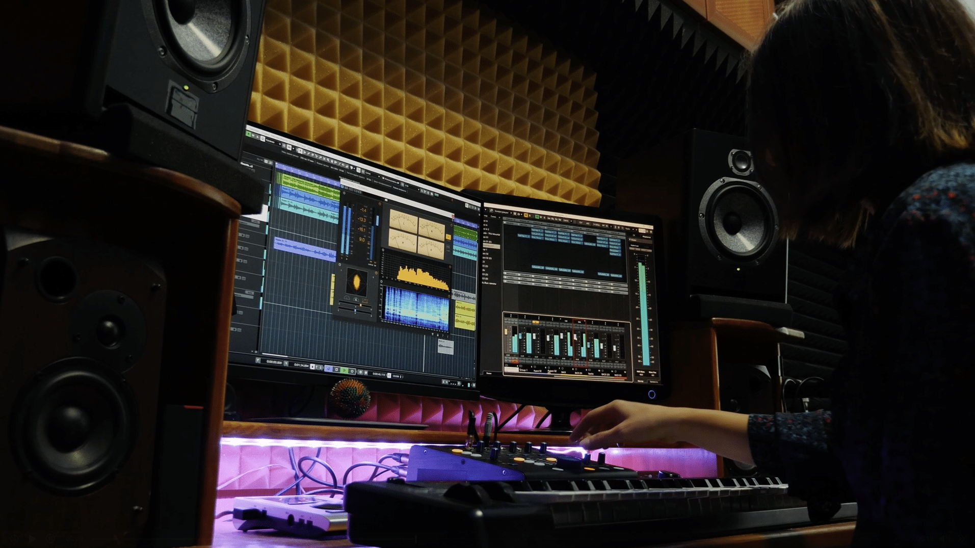Person working at an audio mixing station with two computer monitors displaying sound editing software, surrounded by speakers and soundproofing panels.
