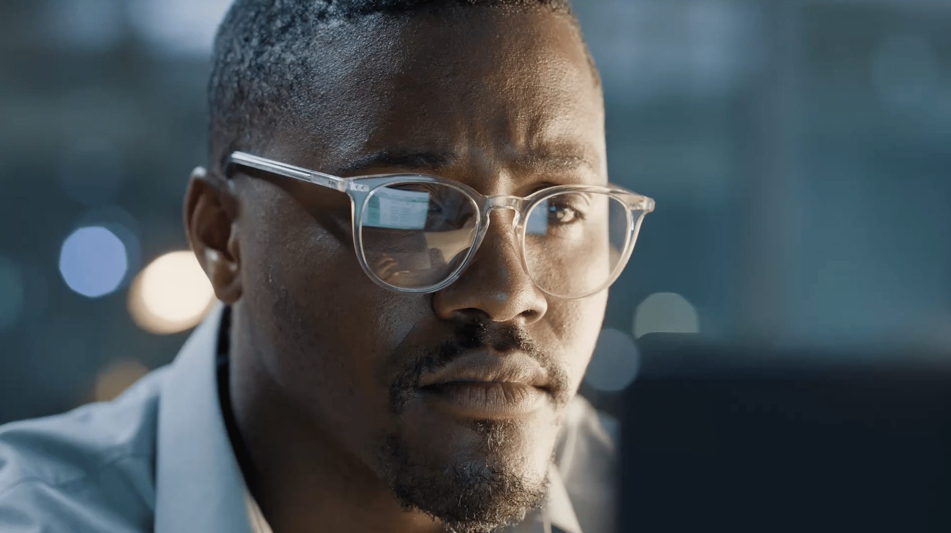 A man with glasses focuses intently on a computer screen, with blurred lights in the background.