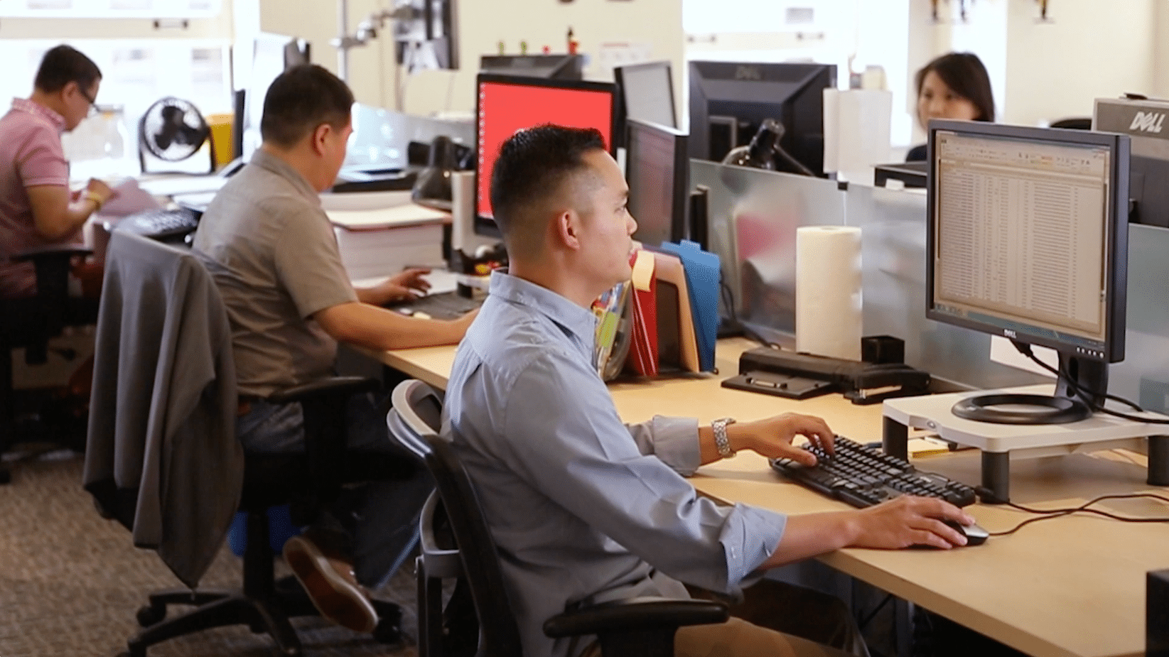 Three people work at computers in a modern office, with desks featuring monitors, keyboards, and office supplies.