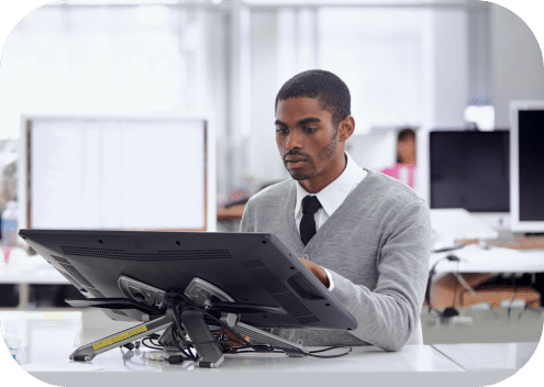 A person in a gray sweater and tie works on a desktop computer in a bright office, diligently compiling customer stories to enhance the company's narrative.