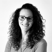Woman with curly hair and glasses, wearing a light-colored top, smiling at the camera against a plain white background.