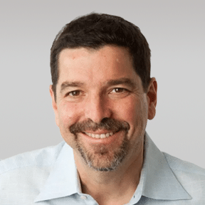 A man with short dark hair, a mustache, and a goatee is smiling. He is wearing a light blue collared shirt against a plain gray background.