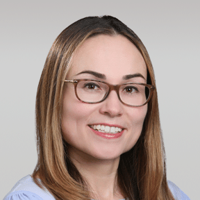 Woman with straight light brown hair and glasses, smiling, wearing a light blue shirt, posed in front of a plain light gray background.