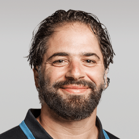 A man with dark hair and a beard smiles at the camera, wearing a black collared shirt with blue trim, against a plain light gray background.