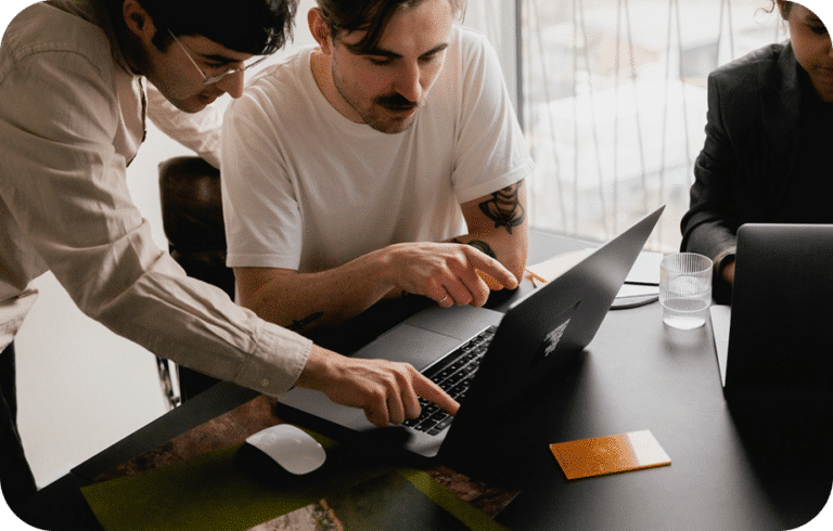 Two people are focused on a laptop, with one pointing at the screen and the other typing. A third person sits nearby. A glass of water and orange card are on the table.