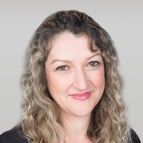 Woman with wavy blonde hair, light skin, and a slight smile, wearing a black top, posing in front of a neutral grey background.