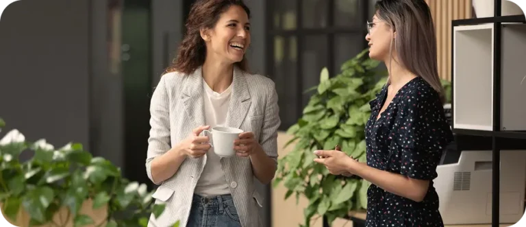 Two women stand indoors near green plants, talking and smiling. One holds a white mug, and both appear to be engaged in a friendly conversation.